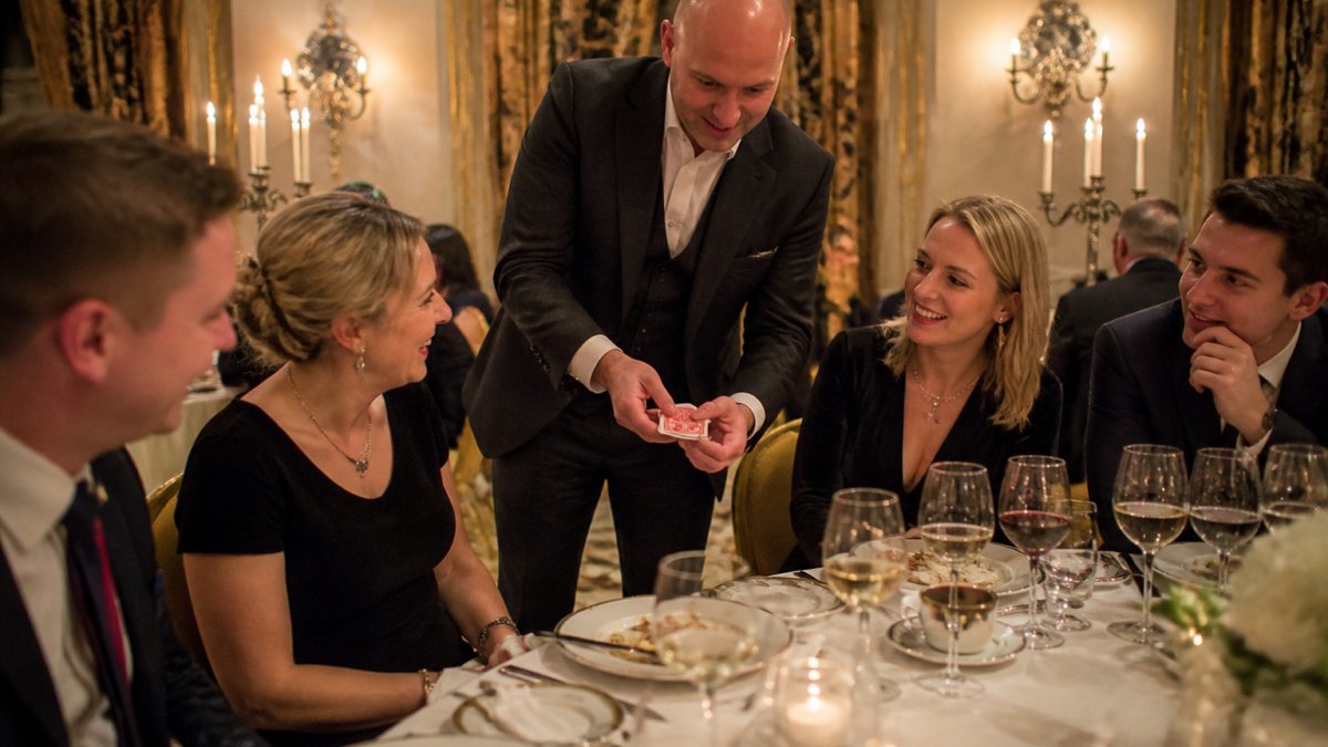 Professional magician performing close-up magic at an elegant hotel dining table with guests seated around, smiling and engaged, with fine china place settings and warm ambient candlelight reflecting off wine glasses