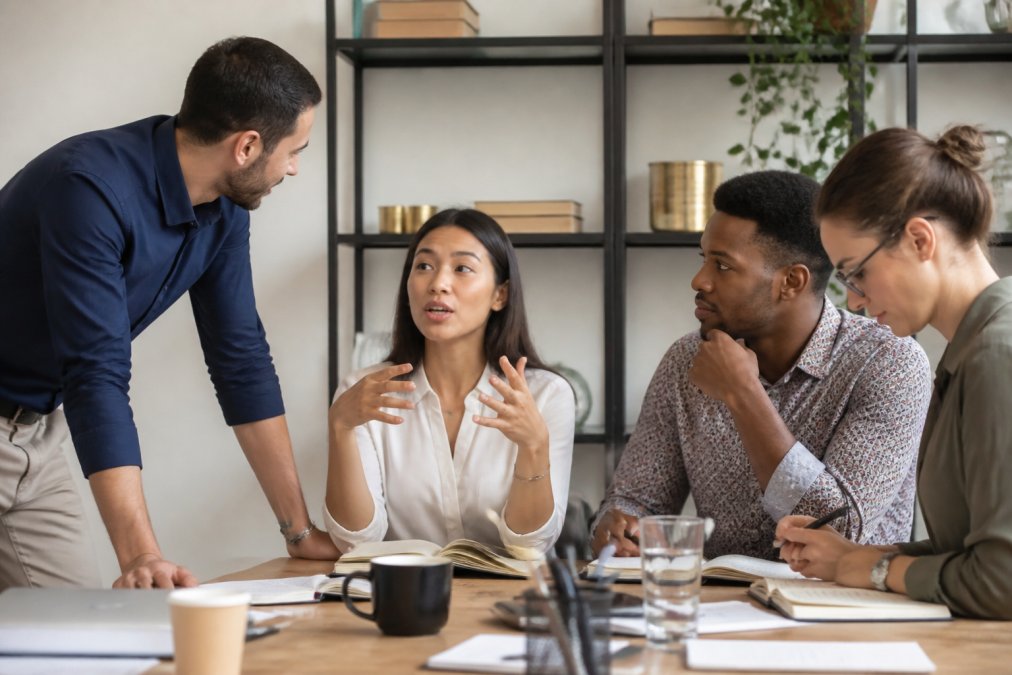 A diverse team of four professionals in a modern office setting, each displaying different body language and engagement styles during a collaborative discussion, one leaning forward confidently, one gesturing animatedly, one listening attentively, and one reviewing detailed notes