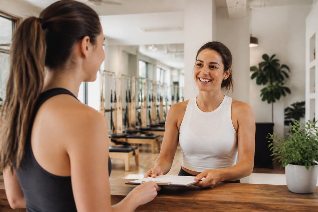 Wellness studio owner or staff member engaging warmly with a new client during a consultation in a modern, well-lit studio space with visible class schedule or equipment in background