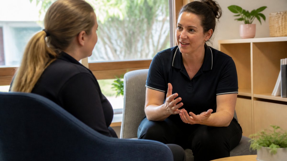 Two professionals sitting face-to-face in a quiet office space, engaged in a calm, respectful conversation. One person is listening attentively while the other speaks. Natural lighting, neutral background, showing body language of openness and psychological safety.