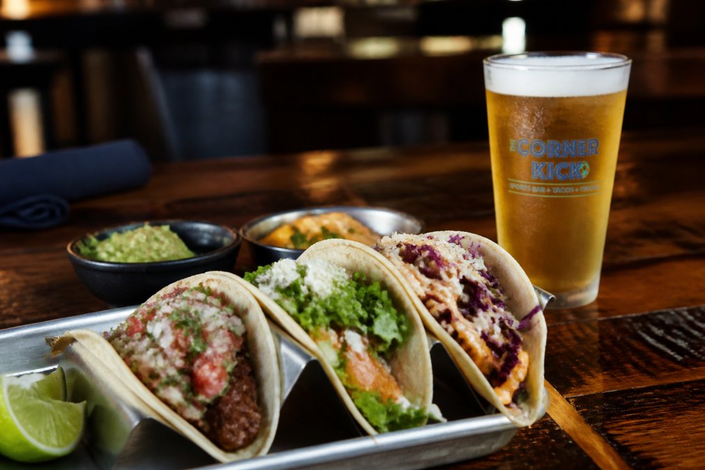 Close-up of scratch-made tacos with colorful toppings arranged on a rustic wooden table, alongside a cold draft beer in a frosted glass, warm ambient lighting in a casual bar-restaurant setting