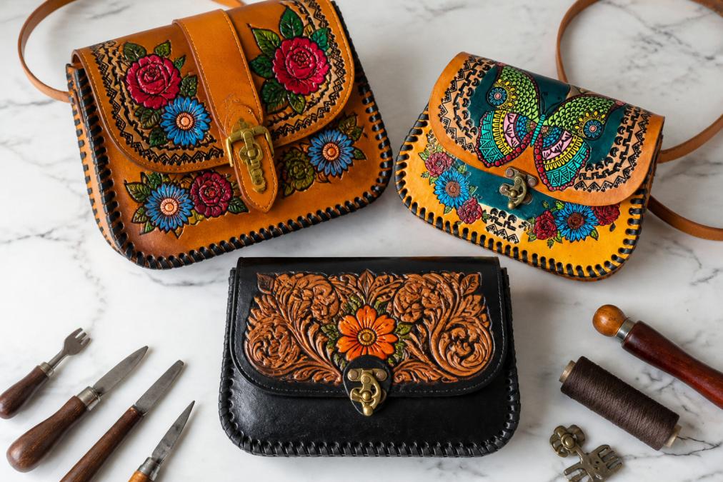 Flat lay of three handcrafted leather handbags with vibrant floral tooling, intricate hand-painted designs, and natural patina displayed on white marble surface with artisan hand tools nearby