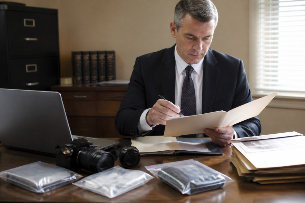 A professional private investigator in a suit examining a case file at a desk with a laptop, evidence bags, and a camera nearby in a well-lit office