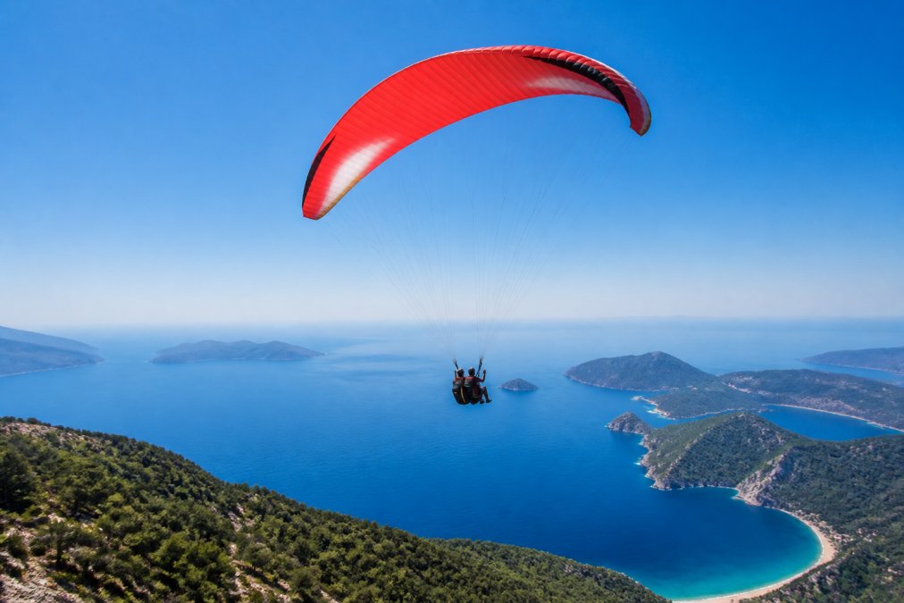 Turist yamaç paraşütü yapıyor, Fethiye'nin mavi denizi ve kıyı manzarası arka planda, güneşli bir gün, gökyüzü açık mavi