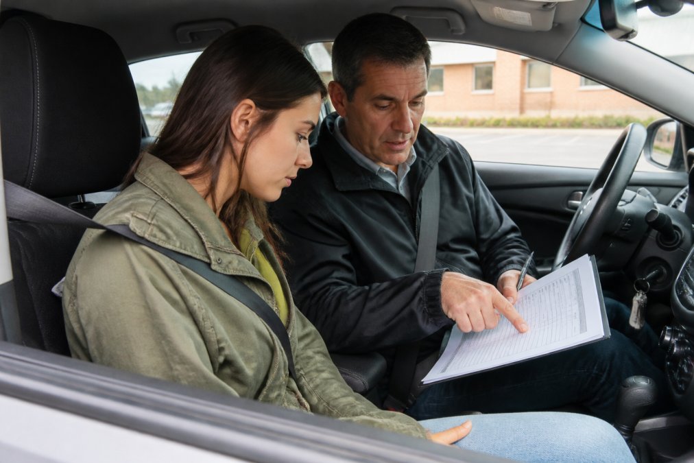 Adult student driver and experienced instructor reviewing a checklist and notes together before a lesson, sitting in a parked vehicle with the instructor pointing to specific items on a printed guide, showing collaborative evaluation and feedback