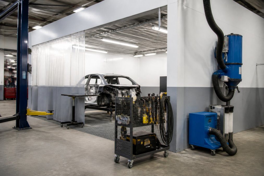 Wide shot of a dedicated aluminum repair bay in a professional body shop, showing a clearly separated workstation with a portable trolley of specialized tools, dust extraction equipment, and bright overhead fluorescent lighting