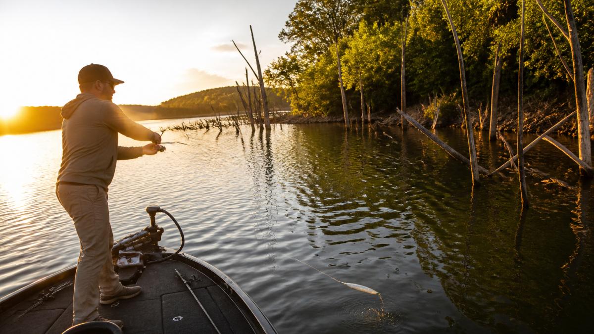 An angler on the front deck of a bass boat at golden hour, casting a glide bait toward submerged timber, with the lure mid-glide visible in the water