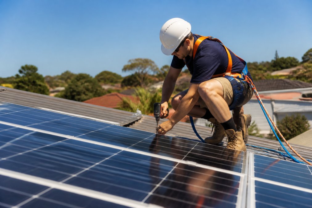 A CEC-accredited solar technician in a high-visibility vest and safety harness crouching beside a rooftop solar panel array on a residential home, checking connections in bright midday sunlight