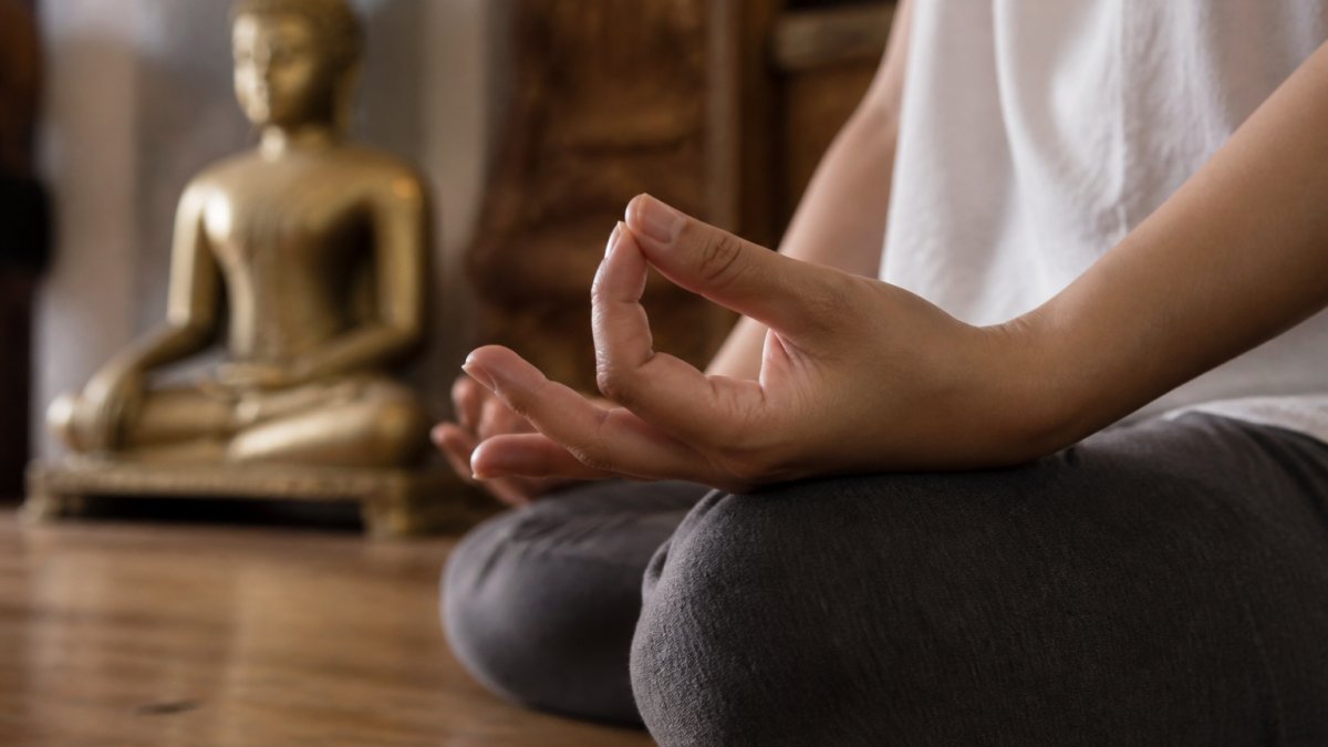 Primo piano delle mani di un praticante che formano un gesto preciso del mudra, appoggiate sulle ginocchia in una sala di meditazione tranquilla e delicatamente illuminata con una statua di Buddha in ottone sfocata sullo sfondo