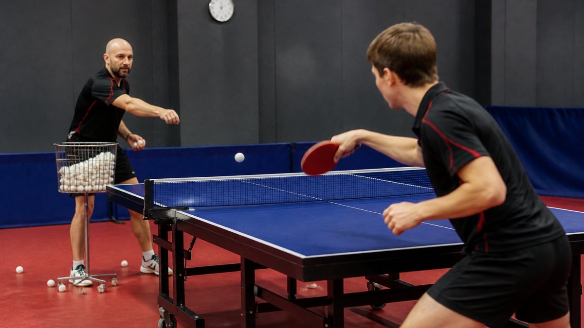 A coach feeding balls rapid-fire from a basket to a focused player mid-forehand stroke in a bright training hall, scattered balls on the floor nearby