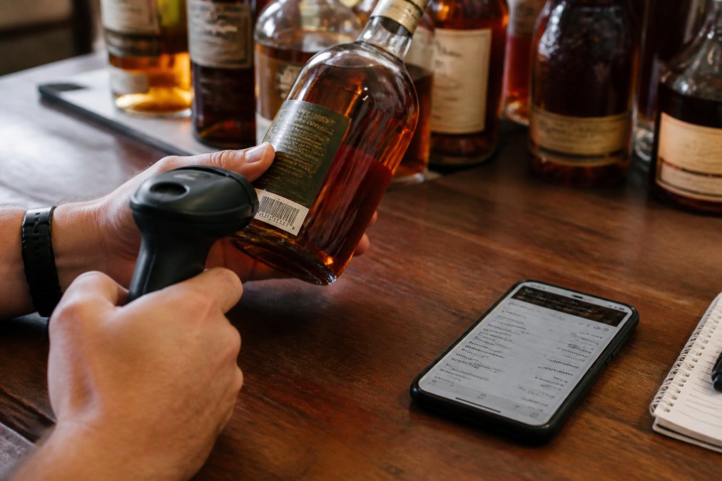 Close-up of hands scanning a whiskey bottle with a barcode scanner, with a smartphone displaying collection data and tasting notes nearby on a wooden table with whiskey bottles in soft background lighting