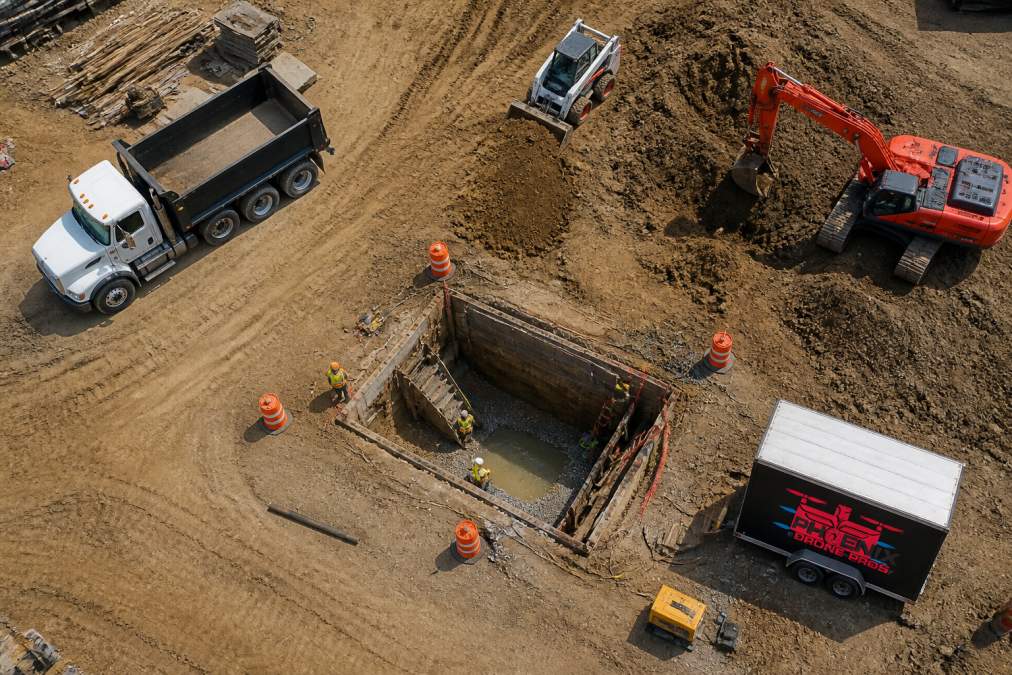 Aerial photograph of active construction site showing workers, heavy equipment, material piles, and potential hazard zones clearly visible from above with bright overhead lighting
