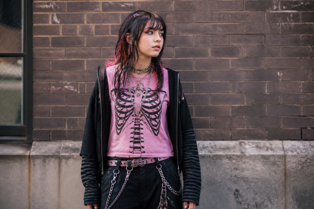 Young person wearing layered Y2K and grunge-inspired streetwear pieces including a pink skeleton graphic tee, vintage accessories, and bold graphic layers against a neutral urban brick backdrop with natural afternoon lighting