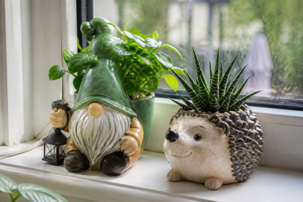 Close-up of character-shaped plant pots with gnome and hedgehog designs with lush green plants, photographed in natural daylight on a windowsill