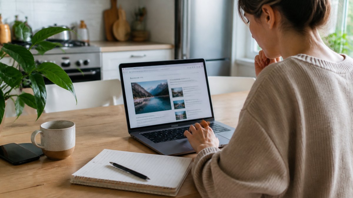 A parent working on a laptop at a kitchen table with a notebook and coffee, reviewing a draft blog post on screen, in a bright home setting