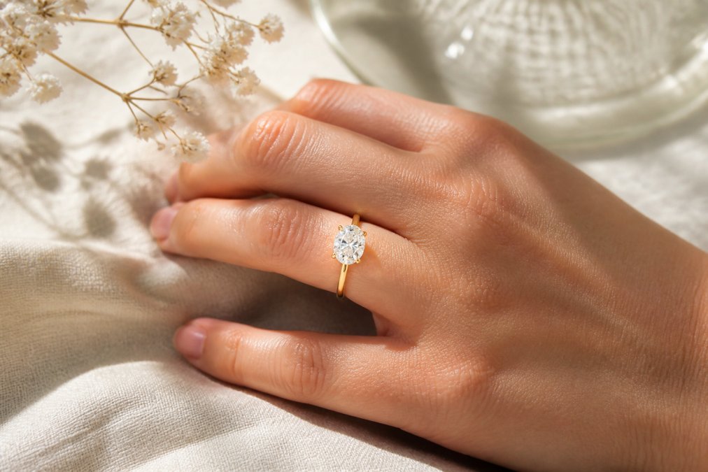 A close-up of a hand wearing a 14k yellow gold solitaire engagement ring with a brilliant oval lab-grown diamond, resting on a soft linen surface with natural window light creating a gentle sparkle
