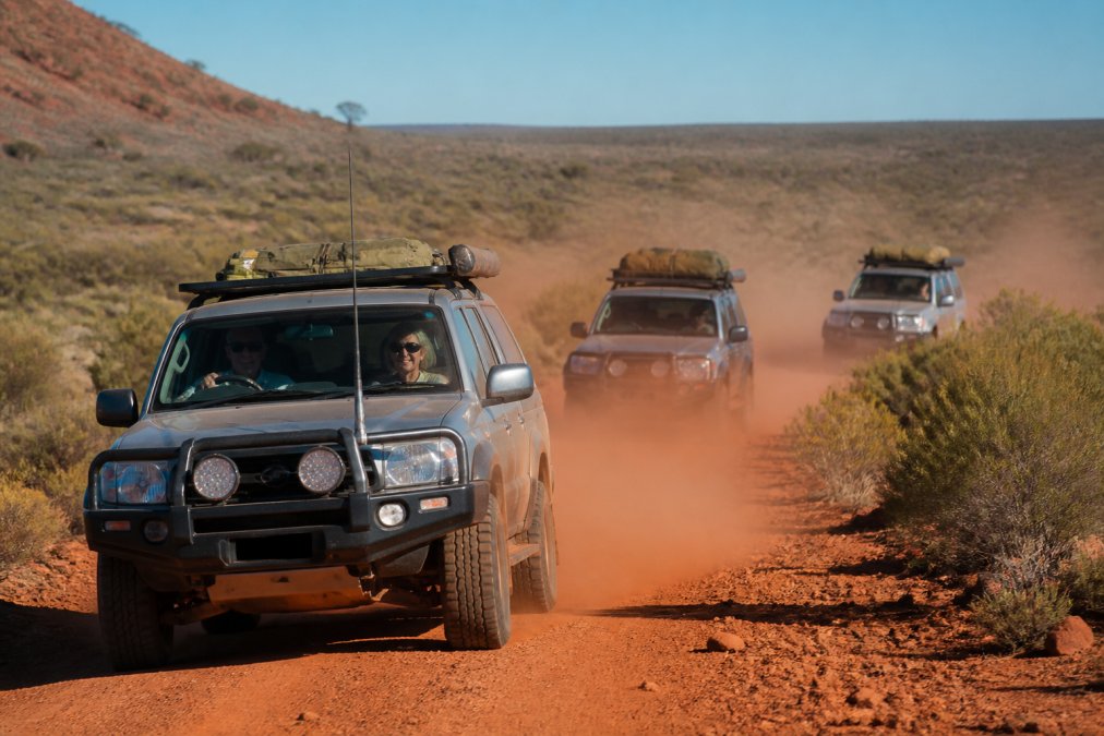 A convoy of three 4WD vehicles kicking up red dust on a remote outback track, with a mature couple in the lead vehicle smiling behind the wheel