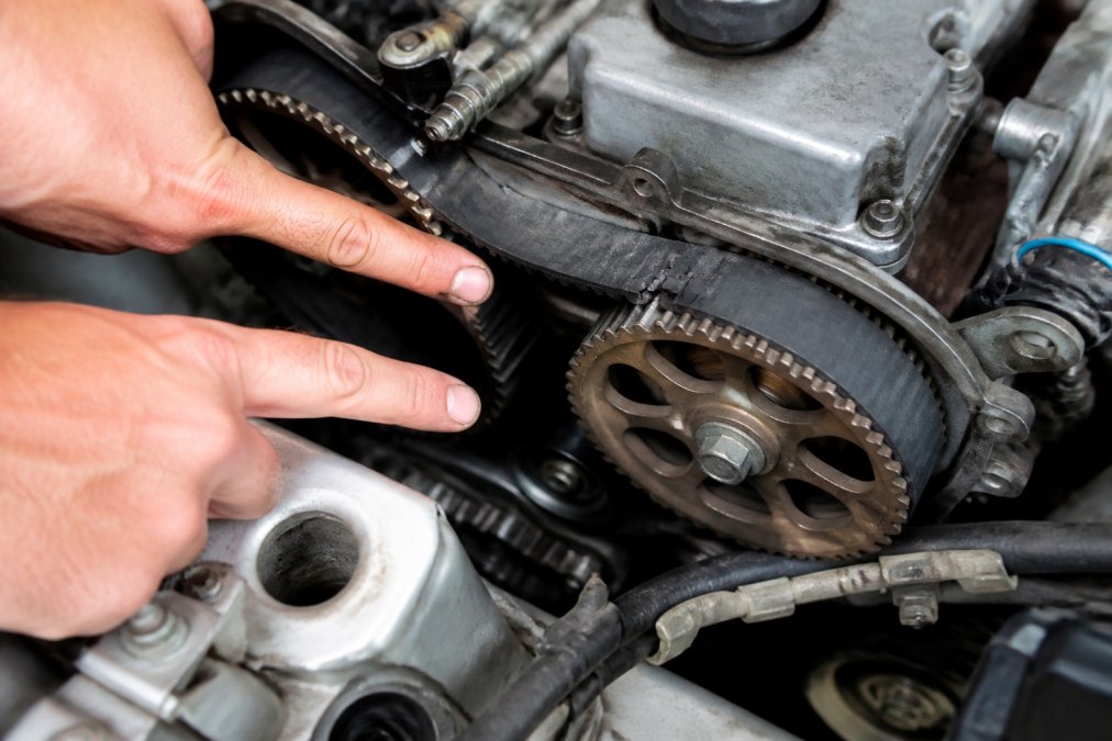 Close-up of an engine bay showing the timing belt area with visible wear patterns, cracks, and fraying on the rubber belt surface, with a technician's hands pointing to deterioration marks