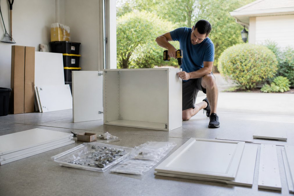 A homeowner assembling a ready-to-assemble kitchen cabinet in a bright garage, cabinet panels and hardware laid out on the concrete floor, cordless drill in hand, natural daylight through an open door