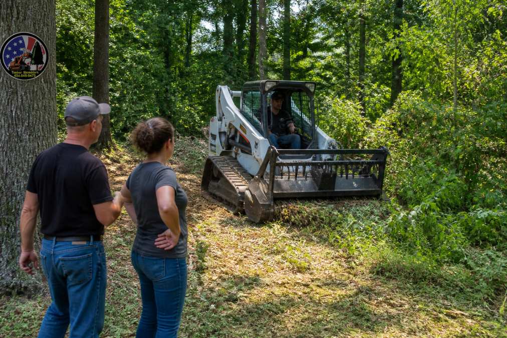 Compact mini skid steer loader with brush cutting attachment clearing overgrown vegetation in a residential property with trees visible in background