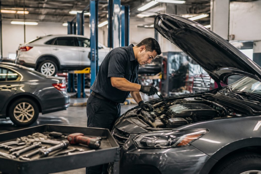 A certified technician in a clean auto repair shop bay performing a vehicle inspection, looking under the hood of a sedan with professional tools laid out on a rolling cart nearby, under bright fluorescent overhead lighting