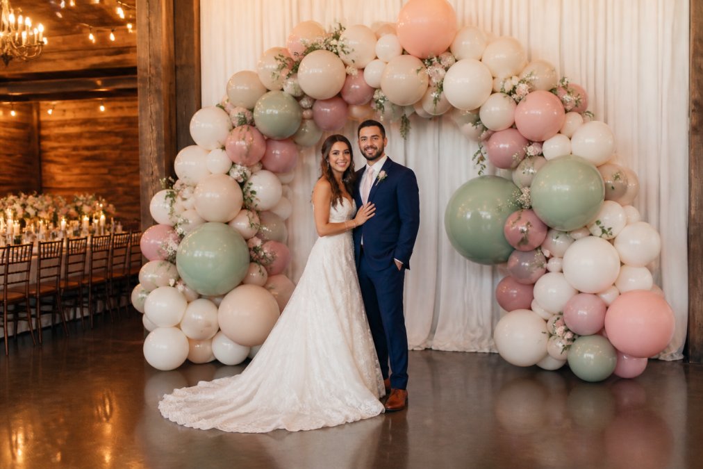 A real-life photo of an elegant wedding reception featuring a large organic balloon arch in matte pastel colors serving as a photo backdrop, with a bride and groom posing in front of it