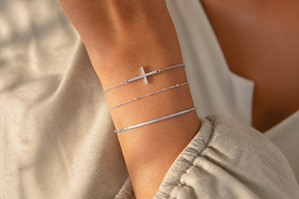 Close-up of a woman's wrist adorned with three delicate bracelets including a sterling silver cross charm bracelet and a thin chain bangle, photographed against a soft natural linen background in warm afternoon light