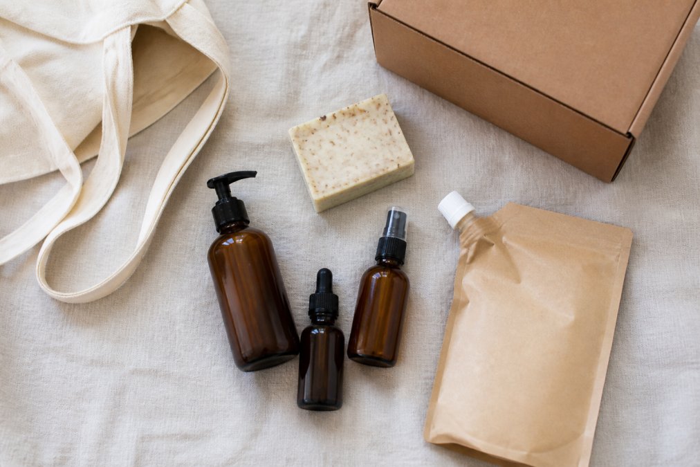 Overhead flat lay of an affordable ethical beauty haul on a pale linen surface: unbranded serum bottles, a bar cleanser, refill pouch, cotton tote and a delivery parcel box, soft natural daylight