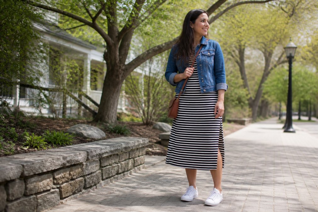 Woman wearing a striped casual dress styled with white sneakers and a denim jacket, standing in natural daylight with trees in background