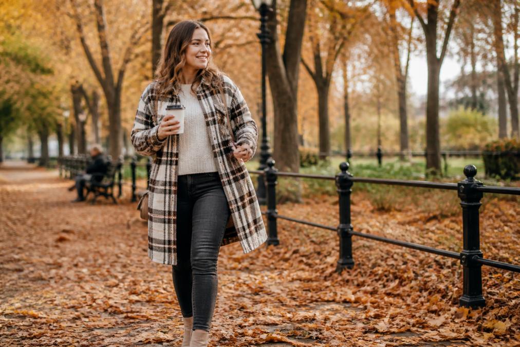 Woman in a plaid shacket and cream sweater walking through a park with autumn leaves while holding a coffee cup