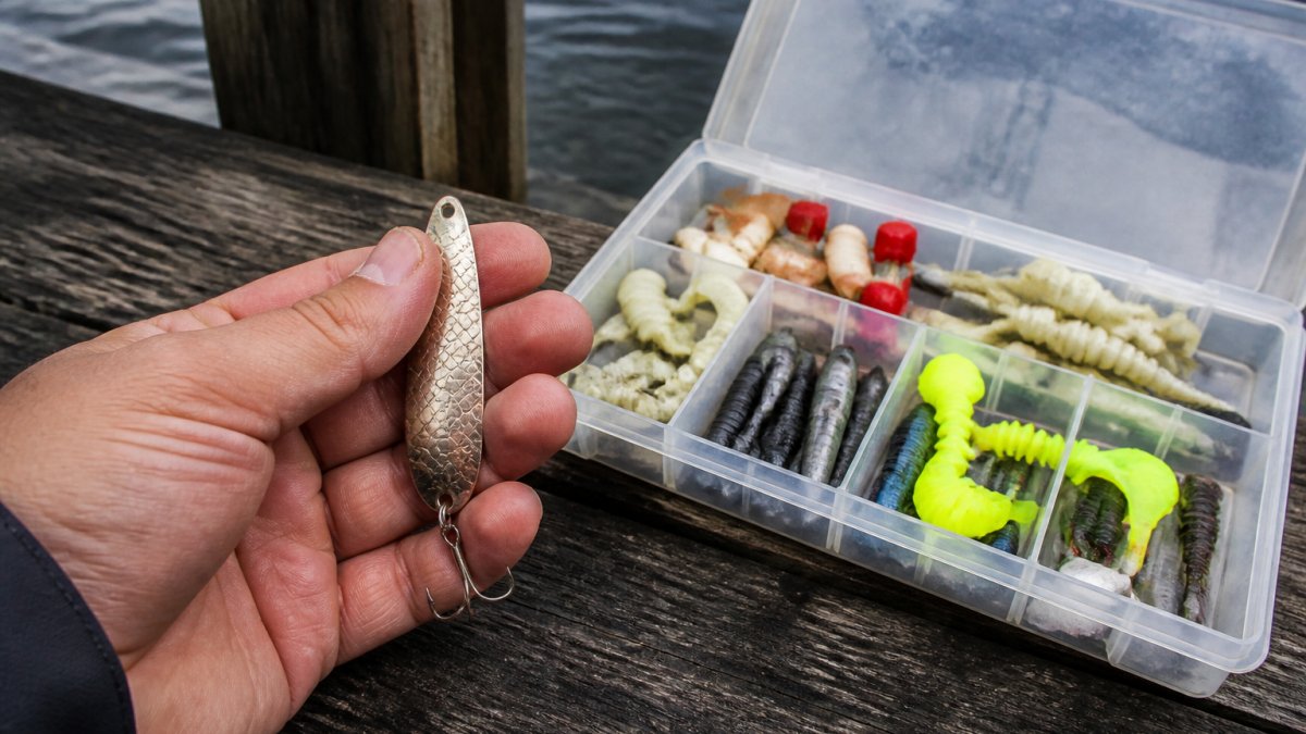 Close-up of a hand holding a metal spoon fishing lure next to an open tackle box filled with poppers and soft grub lures on a timber jetty