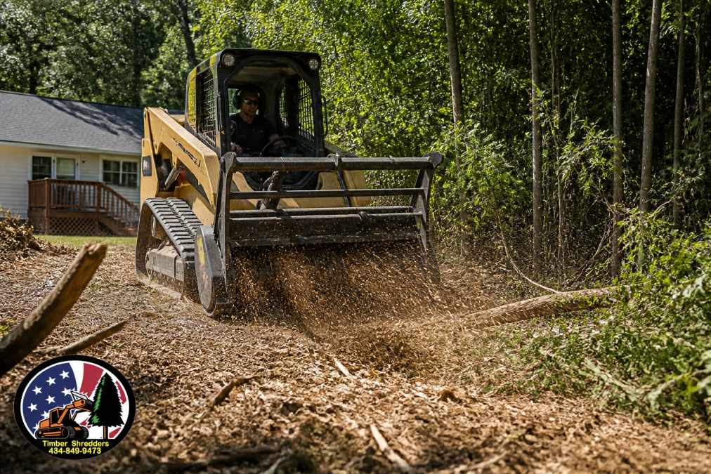 Professional track loader with mulcher head attachment processing dense brush and small trees into fine mulch on an overgrown residential property, with operator visible in cab and mulch material spreading across cleared ground