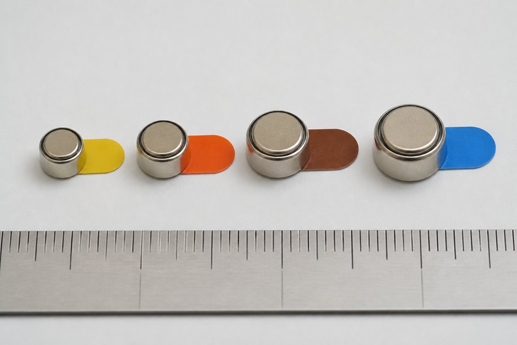 Close-up of four different colored hearing aid batteries (yellow, orange, brown, and blue) arranged in size order on a white surface with a ruler showing scale, color-coded tabs clearly visible