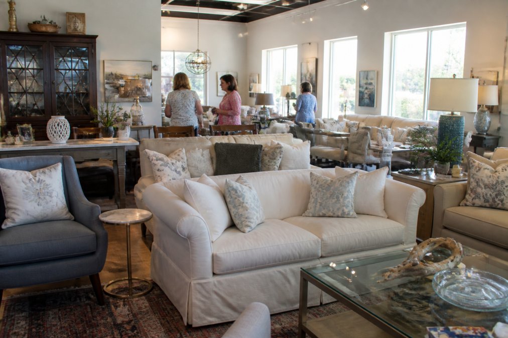 Bright, welcoming consignment store interior with furniture arranged in a 'live in' layout, sofas, chairs, and accent tables displayed as if in a home setting, with natural light streaming through windows and customers browsing pieces