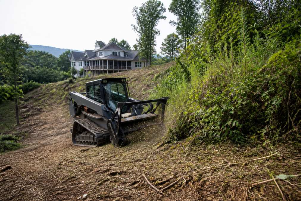 Professional operator using a skid steer loader with forestry mulcher attachment clearing dense vegetation on a sloped property, with mulched material visible on the ground and thick brush being processed