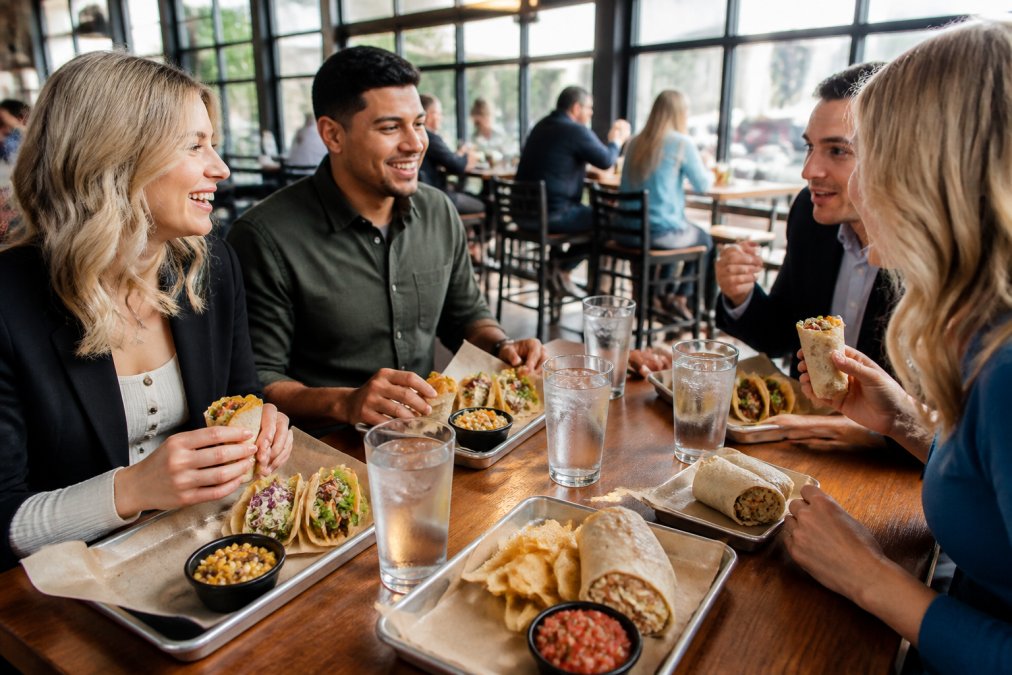 Office workers enjoying a casual lunch break at a vibrant restaurant, gathered around a table with fresh tacos and burritos, natural daylight streaming through windows, relaxed and energized atmosphere