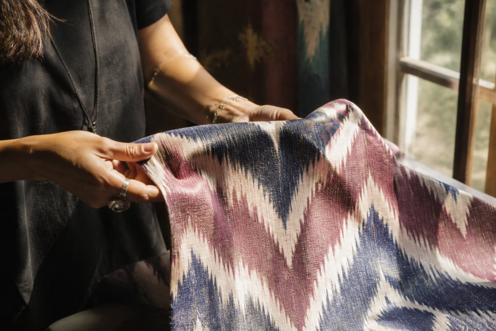 Close-up of a woman's hands turning over an ikat fabric panel, natural window light revealing the woven geometric pattern and the slight irregularity of the dye line