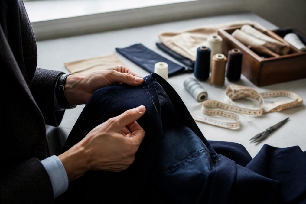 Close-up of hands examining seams and stitching on a luxury garment, fingers tracing the stitchwork and fabric texture against natural window lighting