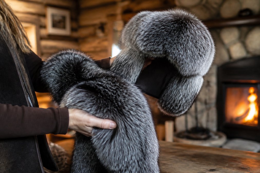 A person's hands gently holding a plush, silver-tipped fur scarf and a matching trapper hat, showing the dense texture and fine craftsmanship against a softly lit, rustic wooden cabin interior with a fireplace glowing in the background