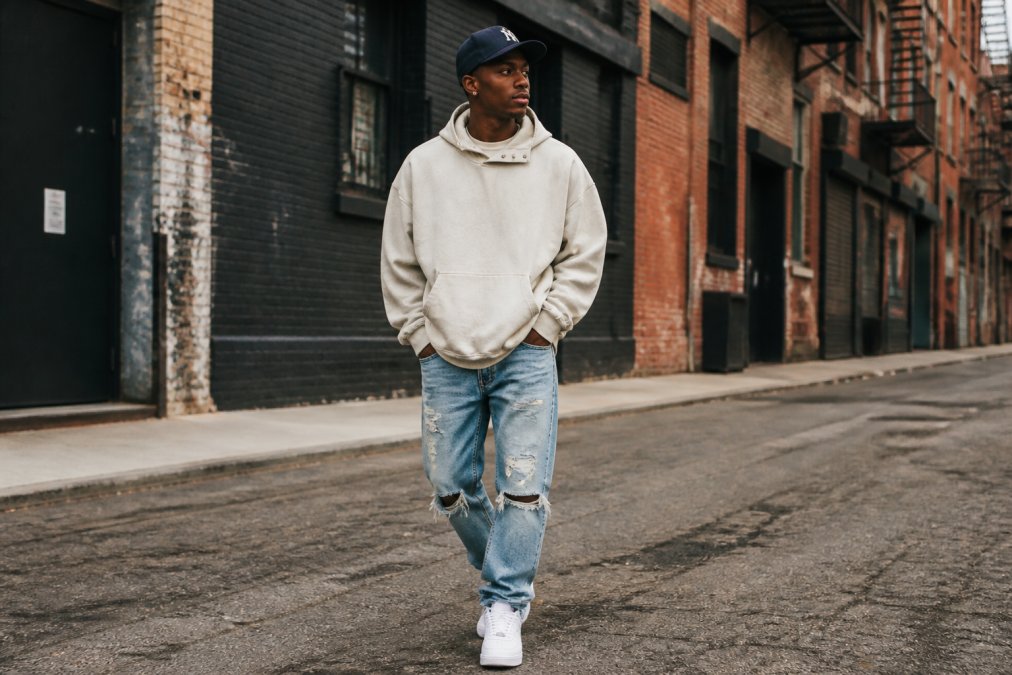 Urban fashion enthusiast wearing an oversized snow-washed snap-collar hoodie in cream with distressed denim and clean white sneakers, posed confidently on a city street with weathered brick buildings and natural daylight