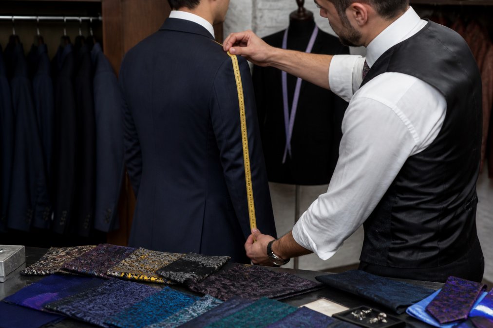 Close-up of a tailor measuring a man's suit jacket shoulder width and sleeve length with a measuring tape, with fabric swatches visible on the workbench
