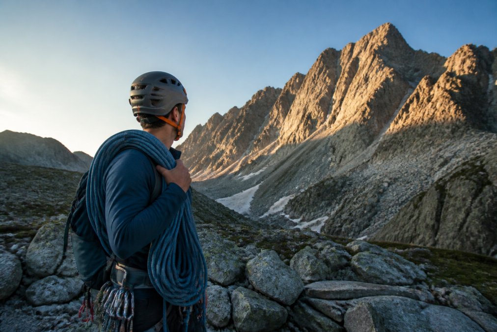 A climber in a helmet and harness, coiled rope over one shoulder, standing at a rocky mountain base in early morning light, studying the ridgeline above