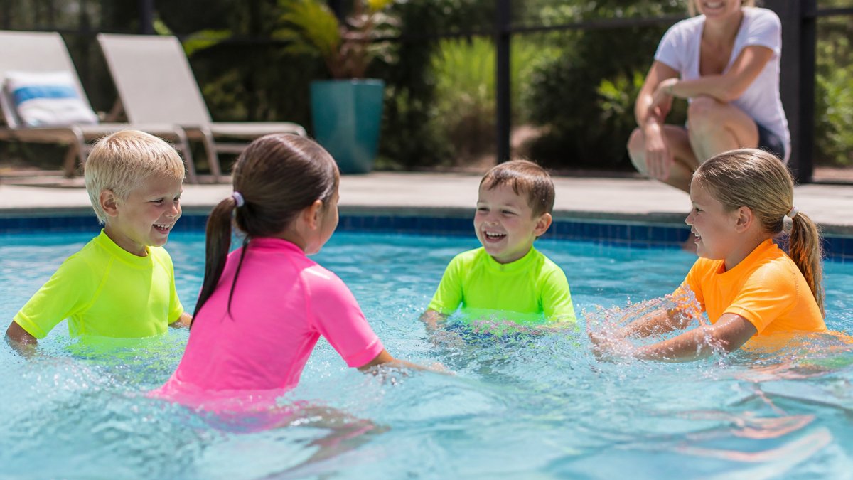 A group of young children in bright neon swimwear playing together in a pool, with a parent watching attentively from poolside in natural afternoon sunlight