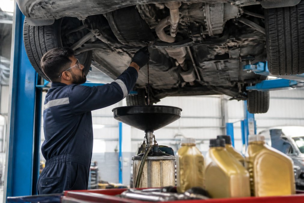 A mechanic in a clean workshop uniform draining engine oil from a car lifted on a hydraulic ramp, workshop tools and amber oil containers visible in the foreground under bright workshop lighting