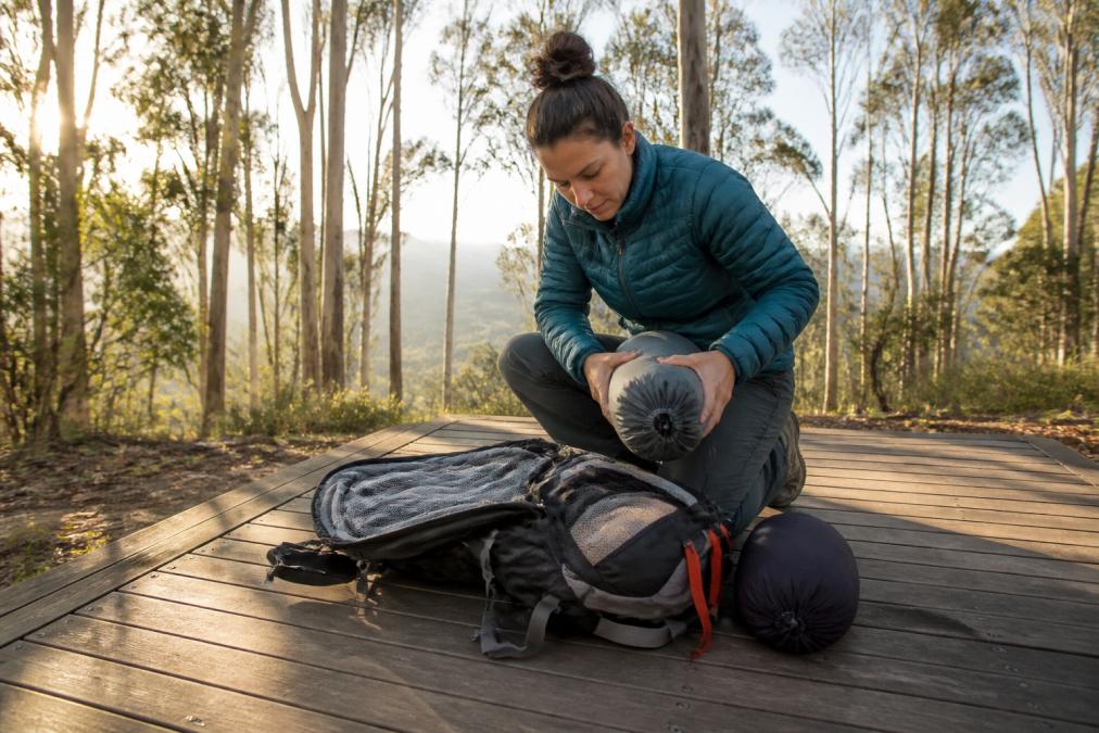 A hiker kneeling beside an open lightweight backpack on a wooden platform, rolling a sleeping bag into its stuff sack, tall eucalypts and soft morning light behind