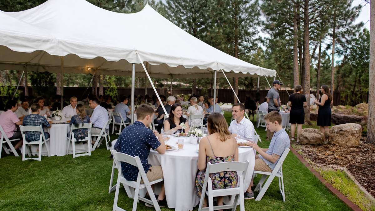 A sunny backyard party setup in Flagstaff with a white event tent, round tables with linens, and folding chairs arranged for guests, pine trees in the background