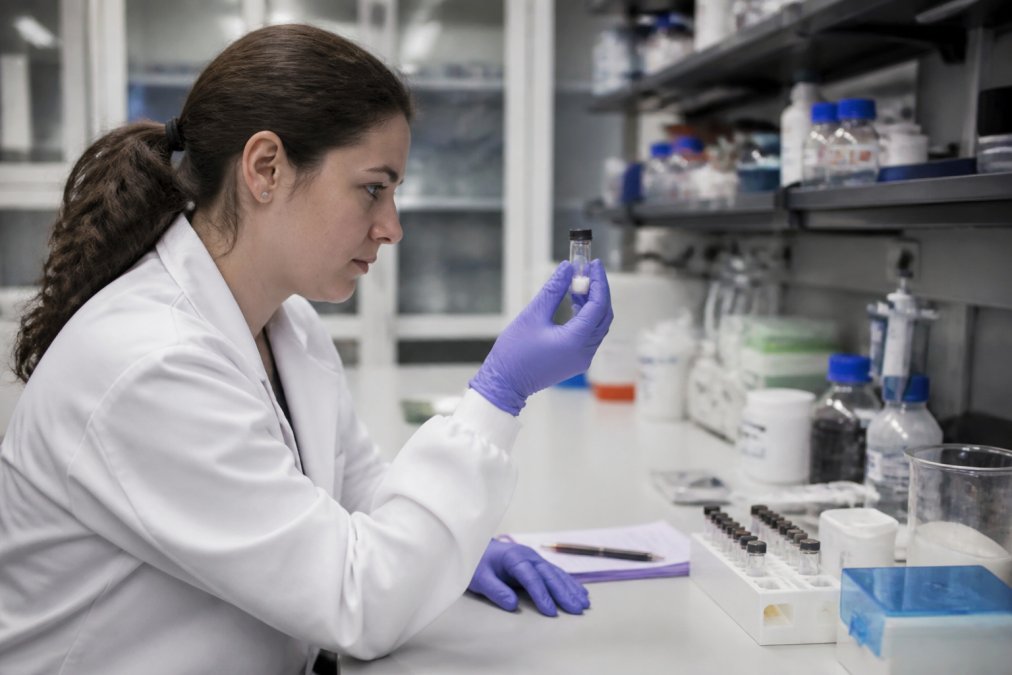 A researcher in a lab coat examining a small vial of lyophilized peptide powder next to a laptop displaying a certificate of analysis document on a clean bench