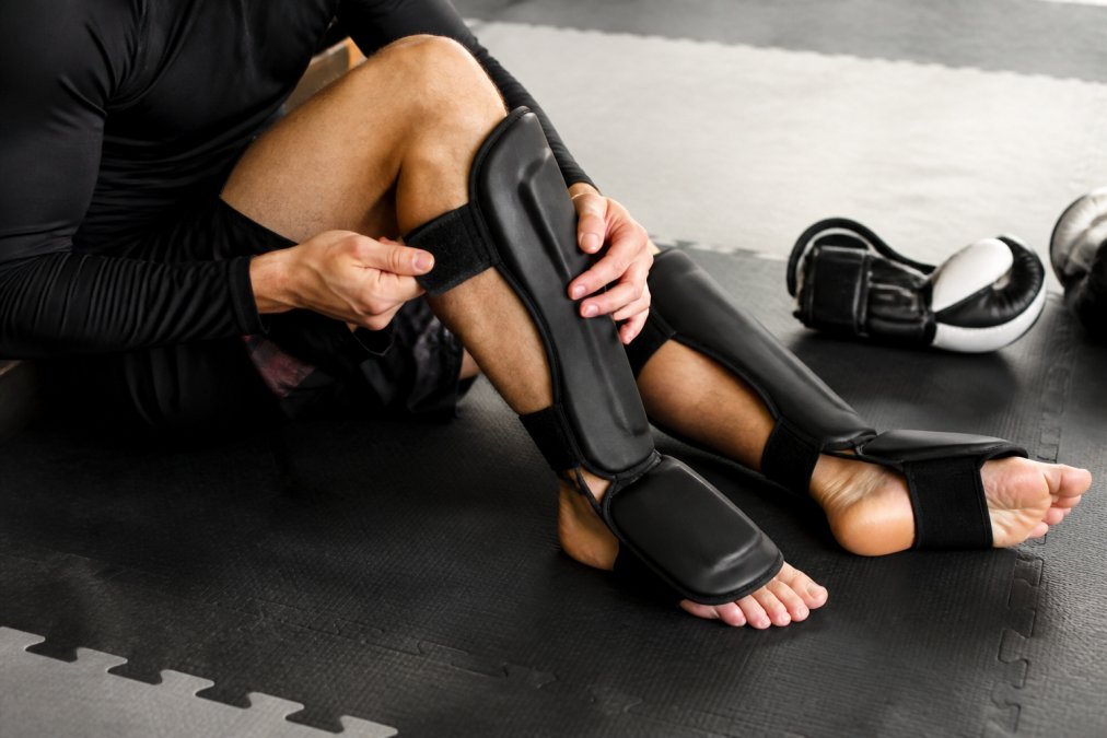 A close-up of a martial artist adjusting the hook-and-loop straps on a pair of black shin guards before sparring on a gym mat