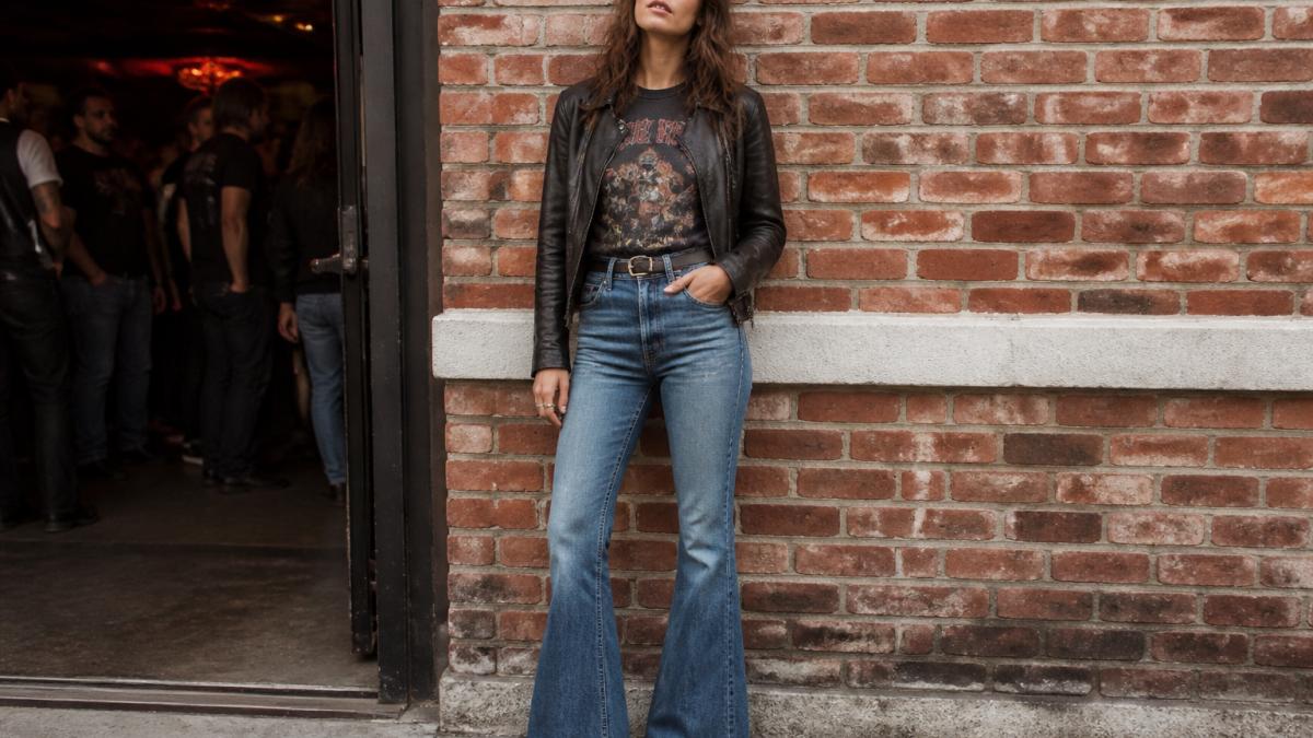 A young adult wearing a vintage graphic band tee tucked into flared jeans with a leather jacket, standing in front of a brick wall at a concert venue, natural lighting