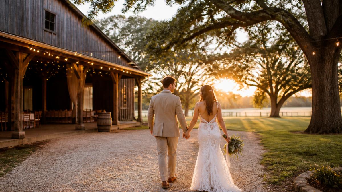 A couple walking hand in hand through a rustic barn venue with string lights and oak trees in the background, golden hour light